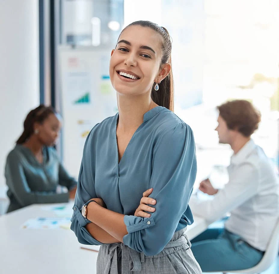 A professional woman in a blue blouse stands with arms crossed, facing a bright office space with colleagues in the background.