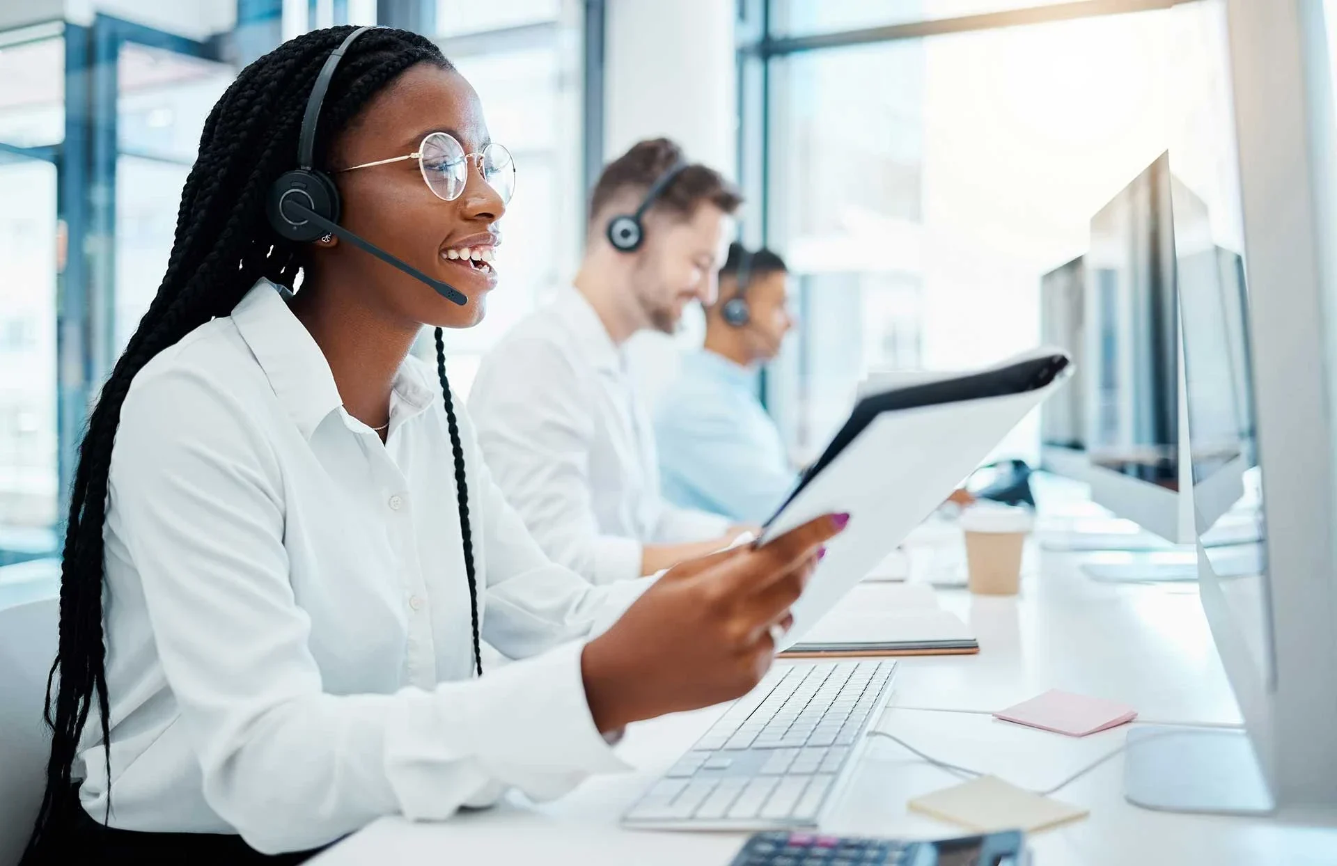 A woman with braided hair wears headphones, holding a clipboard while seated at a modern office desk with computers and colleagues.