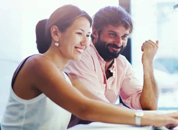 A woman in a tank top and a man in a light pink shirt sit at a table, engaged in conversation, with sunlight coming through a window.